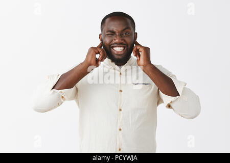 Es ist Gonna blow laut. Portrait von intensiven gestört schöne afrikanische amerikanische Mann im eleganten weißen Hemd mit Brusttasche, Ohren schließen mit Händen, Pressen Stockfoto