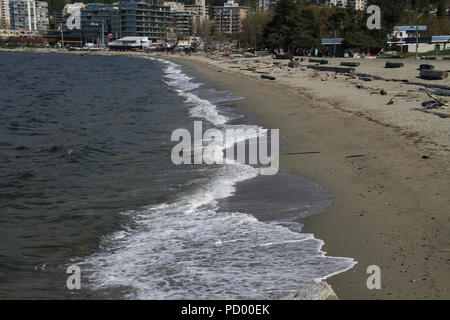 Ein Sandstrand mit einem Stadtbild im Hintergrund. Es gibt Protokolle auf dem oberen Rand des Strandes und kleinen Wellen am Strand. Stockfoto
