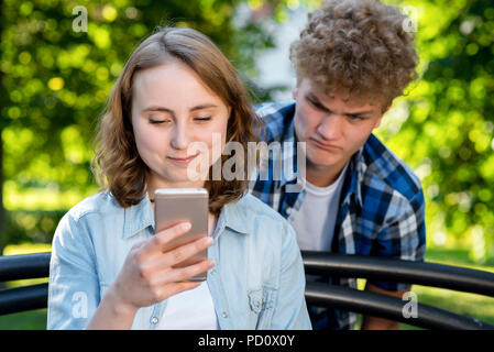 Schöne brünette Mädchen im Sommer in einem Park auf der Natur. In die Hände der Holding ein Smartphone kommuniziert in sozialen Netzwerken. Ein junger Mann Spione auf Ihre Sms. Das Konzept des Misstrauens Problem in der Beziehung. Stockfoto