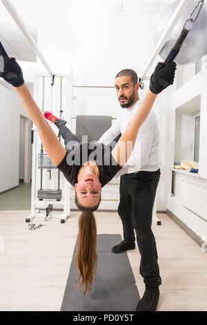 Erwachsene Mann helfen Frau mit Abdominal- Übung wie Sie hält schwarze Gurte an der weißen Metallrahmen befestigt Stockfoto