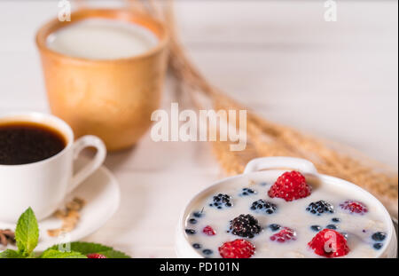 Schale Frühstück Müsli mit Milch und Beeren wie Himbeeren, Heidelbeeren und Brombeeren serviert mit einer Tasse schwarzen Kaffee Stockfoto