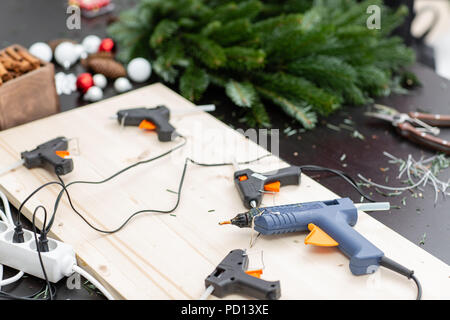 Mehrere Klebepistolen im Vordergrund auf der Platine. Hersteller von Weihnachten Kranz aus Zweigen von Pinien für Urlaub. Master Class auf dem Bilden der dekorativen Ornamenten. Stockfoto