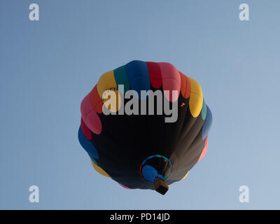 Eine schwarze Heißluftballon mit Rot, Orange, Gelb, Blau, Grün und rosa Akzente auf der Flucht vor einem blauen Himmel. Von unten fotografiert. Stockfoto