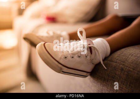 Kleine Mädchen Beine auf Sofa mit schmutzigen weißen Schuhe mit roten Details. Orange Sonnenuntergang aus der Seite. Unscharfer Hintergrund Stockfoto