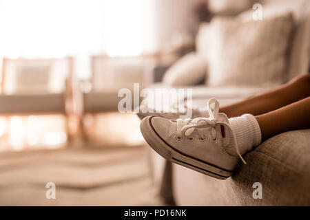 Kleine Mädchen Beine auf Sofa mit schmutzigen weißen Schuhe mit roten Details. Orange Sonnenuntergang aus der Seite. Unscharfer Hintergrund Stockfoto