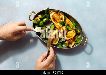 Der Mensch ißt, gebratenen Rosenkohl mit Zucchini in Gusseisen Pfanne serviert auf blauem Stein Gesunde vegetarische Kost Stockfoto