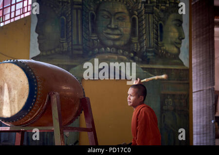 Ein junger buddhistischer Mönch spielen ein tambour während einer Zeremonie an einem Kloster in Siem Reap, Kambodscha, mit Gesichtern der Götter in einem Wandbild in der backgrou lackiert Stockfoto