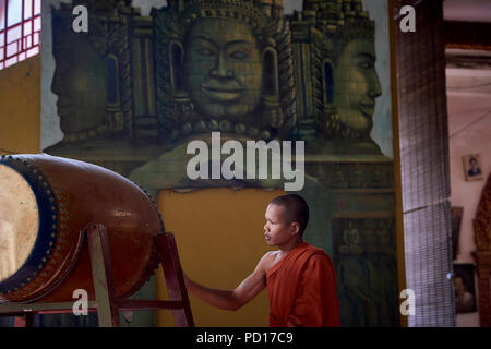 Ein junger buddhistischer Mönch spielen ein tambour während einer Zeremonie an einem Kloster in Siem Reap, Kambodscha, mit Gesichtern der Götter in einem Wandbild in der backgrou lackiert Stockfoto