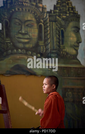 Ein junger buddhistischer Mönch spielen ein tambour, aus dem Rahmen, während einer Zeremonie an einem Kloster in Siem Reap, Kambodscha, mit Gesichtern der Götter in einem Wandbild gemalt Stockfoto