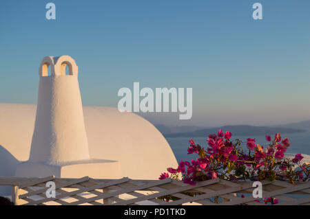 Weiß getünchtes Haus mit Kamin und rosa Blüten mit Blick auf das Meer und den Sonnenuntergang in Oia, Santorini, Kykladen, Griechenland Stockfoto