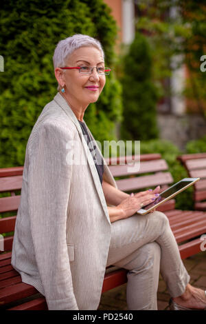 Eine lächelnde Frau ist die Überprüfung einer neuen Tablet während im Park ausruhen. Nahaufnahme Seite Foto anzeigen Stockfoto