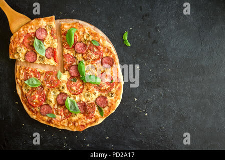 Die Scheiben der Italienische Pizza mit Wurst, Tomaten, Basilikum, Oliven und Käse. Italienische Pizza und Spachtel schließen bis auf schwarzen Hintergrund, Ansicht von oben, Stockfoto