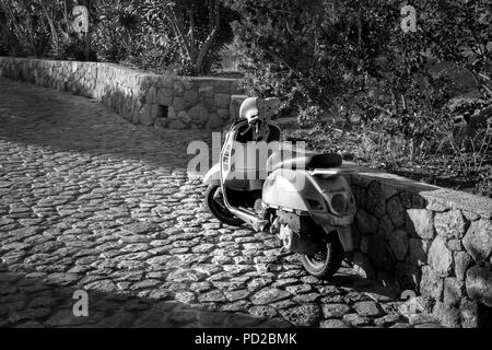 Dies ist ein schwarz-weiß Foto eines Mopeds auf einer gepflasterten Straße geparkt werden. Am Morgen die Sonne auf den Roller scheint. Stockfoto