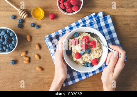 Essen gesundes Frühstück. Haferflocken Porridge mit Himbeeren, Heidelbeeren in Händen. Mädchen essen Hafer für das Frühstück. Ansicht von oben, selektiver Fokus Stockfoto