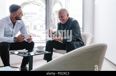 Lächelnd älterer Geschäftsmann mit digitalen Tablet diskutiert mit jungen männlichen Kollege im Büro Lobby. Zwei corporate Professional in Kaffee und casu Stockfoto