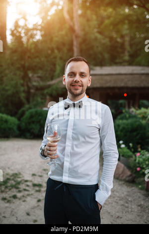 Ein stattlicher Mann in einem weißen Hemd und Krawatte ist Weinprobe Weißwein aus transparentem Glas. Konzept Business Dinner. Outdoor Portrait. Stockfoto