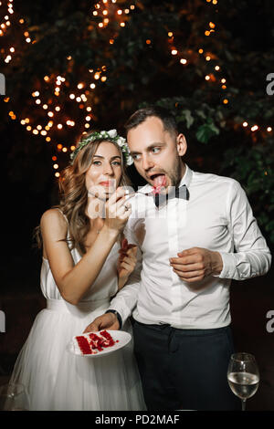 Glückliche Braut und Bräutigam die Hochzeitstorte in der vorderen Girlande Licht Dekoration. Hochzeit Abend im Freien. Süße Happy Life Konzept. Stockfoto