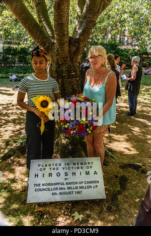 August 6, 2018, London, UK. 6. August 2018. Frauen halten eine Sonnenblume und den Kranz vom Bürgermeister von Camdent vor der Hiroshima Kirschbaum am Ende der London CND Zeremonie im Gedenken an die Opfer der Vergangenheit und Gegenwart auf der 73. Jahrestag der Abwurf der Atombombe auf Hiroshima, auch das Erinnern an die getötet und das Leben mit den Auswirkungen der Strahlung, die durch die zweite Atombombe auf Nagasaki drei Tage später. Die Beiträge enthalten eine Lesung in englischer Sprache der Erklärung früh am Tag an der Gedenkfeier in Hiroshima, und ein Appell von Rebecca Johnson, auf der Stockfoto