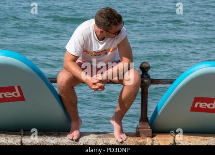 Cowes, Großbritannien. 6. August 2018. Die hohe tempertatures weiter und die Leute machen das Beste aus der heißen suuny Wetter in Cowes auf der Isle of Wight. Ein junger Mann mit zwei Paddle Boards sitzen auf dem Meer Wand. paddleboards Menschen in der Sonne beobachten die Segelyacht rennen und Zeit miteinander zu verbringen am Strand im heißen August Sonnenschein. Quelle: Steve Hawkins Fotografie/Alamy leben Nachrichten Stockfoto