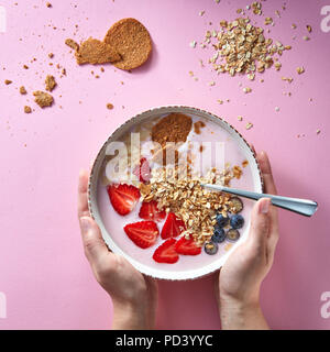 Woman's Hände halten eine Schüssel mit organischen Joghurt Smoothie mit Erdbeeren, Banane, Blaubeere, Haferflocken und Kekse auf rosa Hintergrund. Flach. Stockfoto