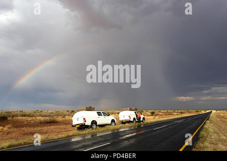 Touristische Autos 4x4 durch gepflasterte Straße in stürmischen Wetterbedingungen mit einem Regenbogen über der Straße geparkt, Namibia Stockfoto