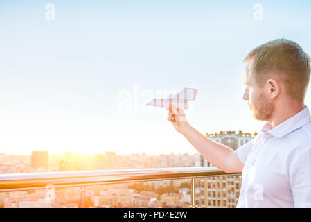 Der Mann hält Papierflieger in blauer Himmel bei Sonnenuntergang Licht mit Stadt Landschaft Hintergrund. Träume und Freiheit Konzept. Selektive konzentrieren. Copyspace Stockfoto