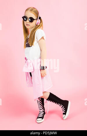 Mädchen mit Red Tails auf einem rosa Hintergrund. Ein bezauberndes Mädchen in heller Kleidung, Sonnenbrille und Kopfhörer auf ihren Fuß zurück. Stockfoto