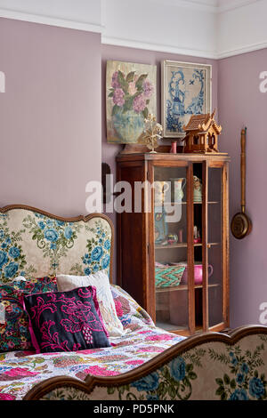 Französische Bettgestell gepolstert in Vintage geblümten Stoffen im Schlafzimmer in einer staubigen Violett Farbe lackiert. Die Glasfront display cabinet Edwardian ist. Stockfoto