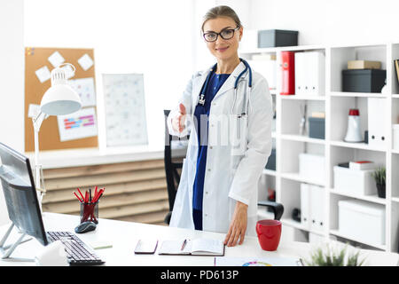 Ein junges Mädchen in einem weißen Gewand steht in der Nähe der Schreibtisch im Büro und erstreckt sich ihre rechte Hand nach vorn. Ein Stethoskop um den Hals hängt. Stockfoto