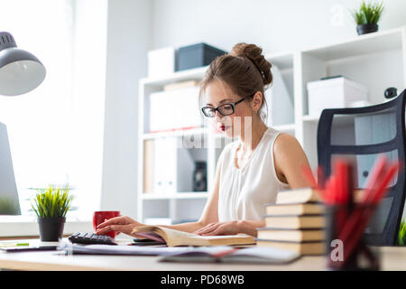 Ein junges Mädchen sitzt an einem Computer Tisch und hält ein aufgeschlagenes Buch in den Händen und druckt auf der Tastatur. Stockfoto