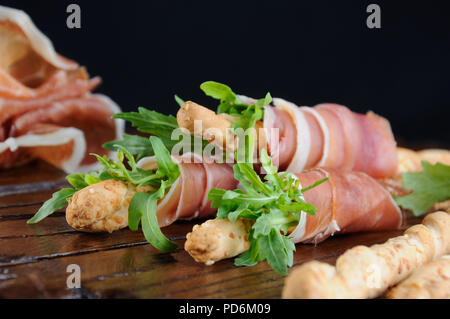 Grissini - Brot Sticks mit Parmesan, umwickelt mit einem Stück Schinken und Rucola. Italienische Gericht mit antipast auf einem Holztisch. Stockfoto