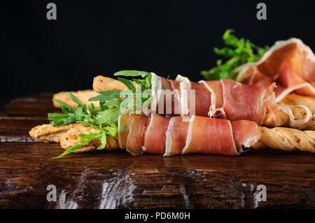 Grissini - Brot Sticks mit Parmesan, umwickelt mit einem Stück Schinken und Rucola. Italienische Gericht mit antipast auf einem Holztisch. Stockfoto