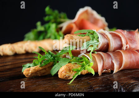 Grissini - Brot Sticks mit Parmesan, umwickelt mit einem Stück Schinken und Rucola. Italienische Gericht mit antipast auf einem Holztisch. Stockfoto