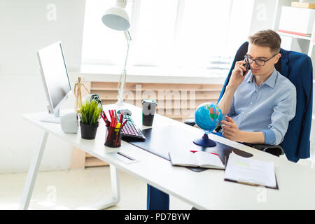 Ein junger Mann sitzt im Büro am Computer, am Telefon zu sprechen und auf der ganzen Welt. Stockfoto
