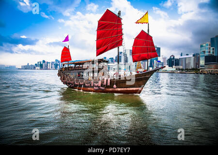 Traditionelle Chinesische Holz Segelschiff mit Red Sails in Victoria Harbour in den Strahlen der untergehenden Sonne. Wolkenkratzer in der Innenstadt von Hong Kong sind sichtbar Fro Stockfoto