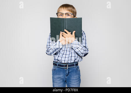 Eine charmante junge mit einem eingelegten Hemd und Jeans steht auf einem grauen Hintergrund. Der junge geschlossen sein Gesicht Buch Stockfoto