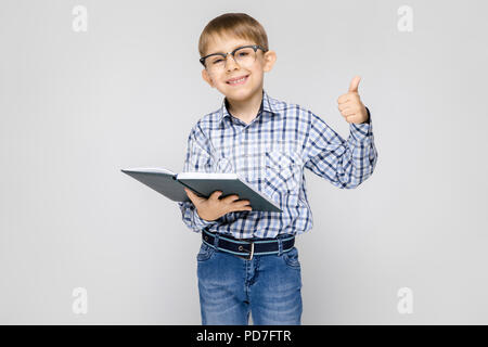 Eine charmante junge mit einem eingelegten Hemd und Jeans steht auf einem grauen Hintergrund. Der Junge hält ein Buch in der Hand. Junge mit Brille Stockfoto