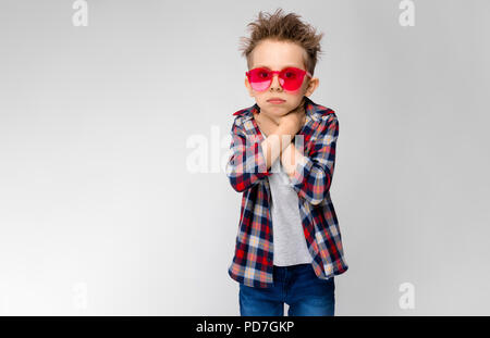 Ein schöner Junge in einem Plaid Shirt, grau Shirt und Jeans steht auf einem grauen Hintergrund. Ein Junge in rot Sonnenbrille. Der Junge hält seine Hände in seine Kehle. Stockfoto