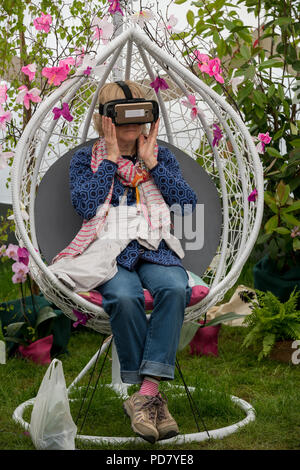 Frau mit VR-Headset, setzen Sie sich unter den Blumen an der Virtuellen Realität orchid Erfahrung - RHS Chatsworth Flower Show, Derbyshire, England, UK. Stockfoto