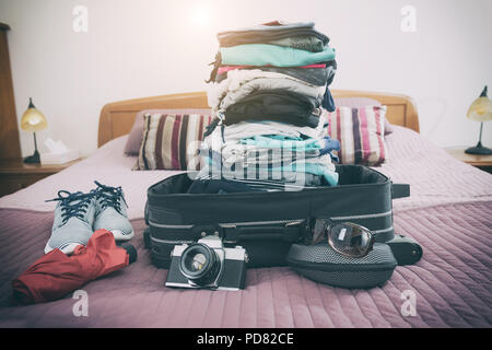 Gepäck mit Stapel von Kleidung, Kamera, Sonnenbrille und andere Elemente auf dem Bett Stockfoto