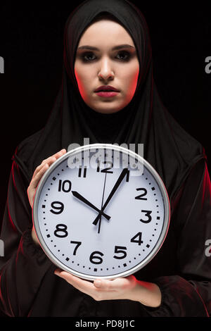 Porträt der schönen schweren Angst junge muslimische Frau mit schwarzen Hijab halten Sie clock in ihre Hände als Laufzeit Konzept auf schwarzem Hintergrund Angst Stockfoto