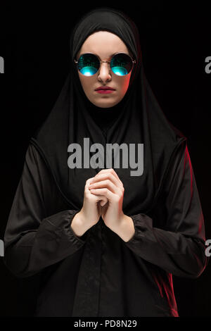 Portrait von schöne stilvolle junge muslimische Frau mit schwarzen Kopftuch und einer Sonnenbrille als moderne östlichen Fashion Concept auf schwarzem Hintergrund Stockfoto