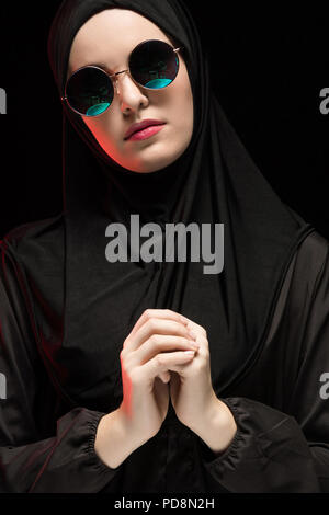 Portrait von schöne stilvolle junge muslimische Frau mit schwarzen Kopftuch und einer Sonnenbrille als moderne östlichen Fashion Concept auf schwarzem Hintergrund Stockfoto