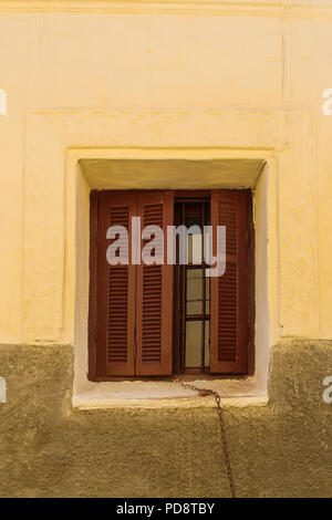 Gelbe Wand eines Hauses mit einem Fenster mit teilweise geöffnet braun Shutter. Kette hängen vom Fenster verrostet. El Jadida, Marokko. Stockfoto