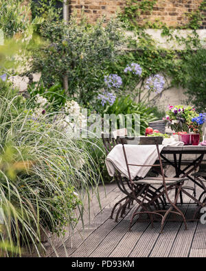 Alte französische Bistro Stühle um langen Holztisch auf Garten Dachterrasse mit Belag. Agapanthus und Jasmin blüht im Hintergrund Stockfoto