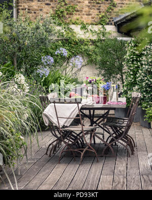 Alte französische Bistro Stühle um langen Holztisch auf Garten Dachterrasse mit Belag. Agapanthus und Jasmin blüht im Hintergrund Stockfoto