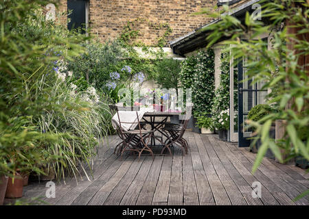 Alte französische Bistro Stühle um langen Holztisch auf Garten Dachterrasse mit Belag. Agapanthus und Jasmin blüht im Hintergrund Stockfoto