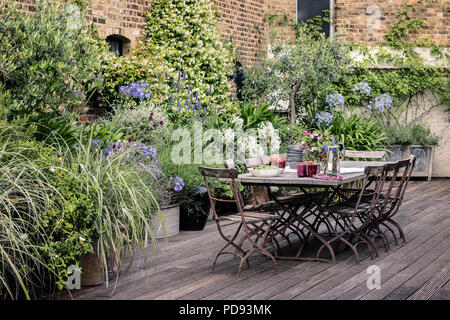 Alte französische Bistro Stühle um langen Holztisch auf Garten Dachterrasse mit Belag. Agapanthus und Jasmin blüht im Hintergrund Stockfoto