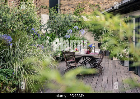 Alte französische Bistro Stühle um langen Holztisch auf Garten Dachterrasse mit Belag. Agapanthus und Jasmin blüht im Hintergrund Stockfoto