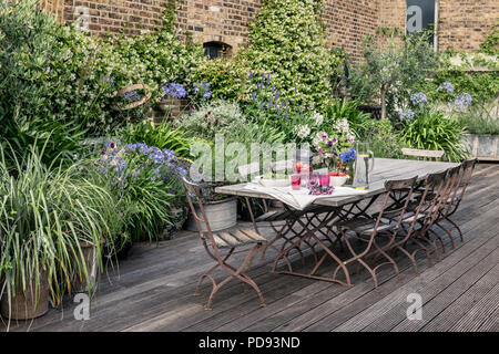 Alte französische Bistro Stühle um langen Holztisch auf Garten Dachterrasse mit Belag. Agapanthus und Jasmin blüht im Hintergrund Stockfoto